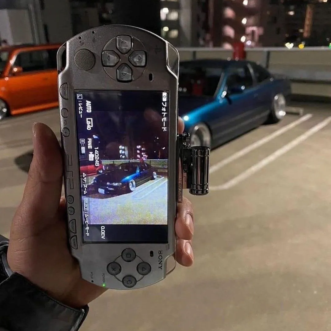 A hand holding a silver PSP-1000 with a camera accessory mounted on top, photographing a modified car in a parking garage at night.