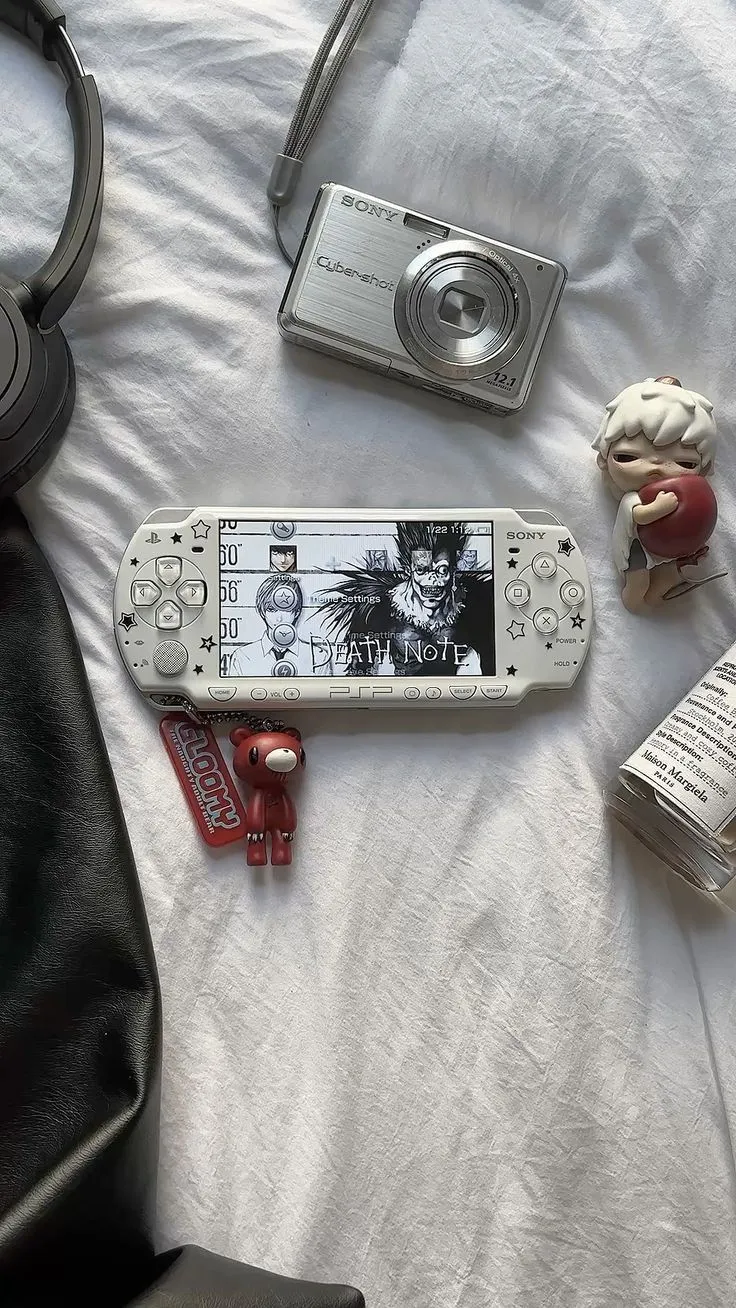 A white PSP running a Death Note themed screen, styled on a bed next to a Sony Cybershot digital camera, a red bear keychain, and a Maison Margiela fragrance bottle.