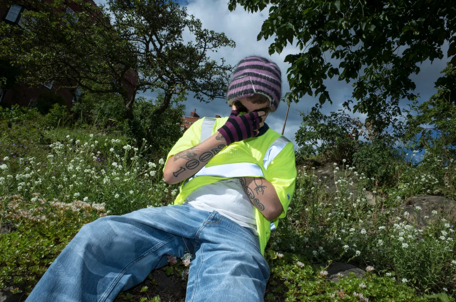 Yung Lean in a neon safety vest and striped beanie, seated in an overgrown garden.