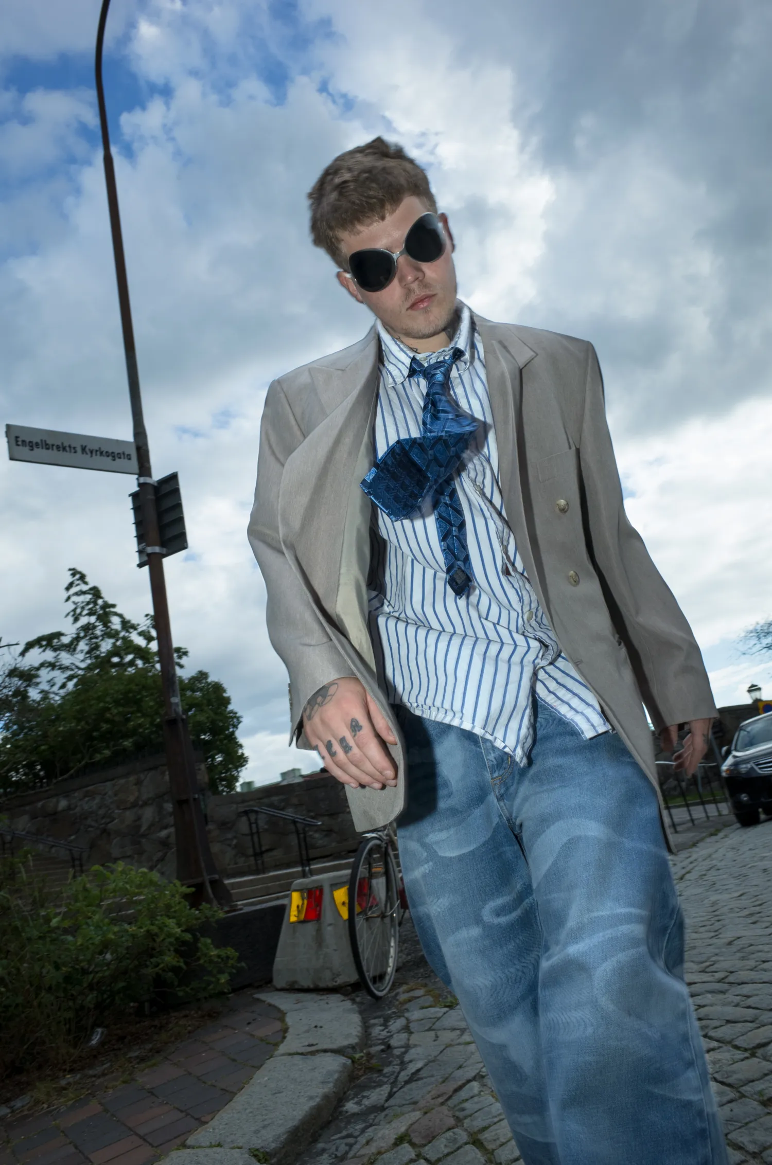 Yung Lean in a beige blazer and unravelling striped tie, walking down a cobbled Stockholm street.