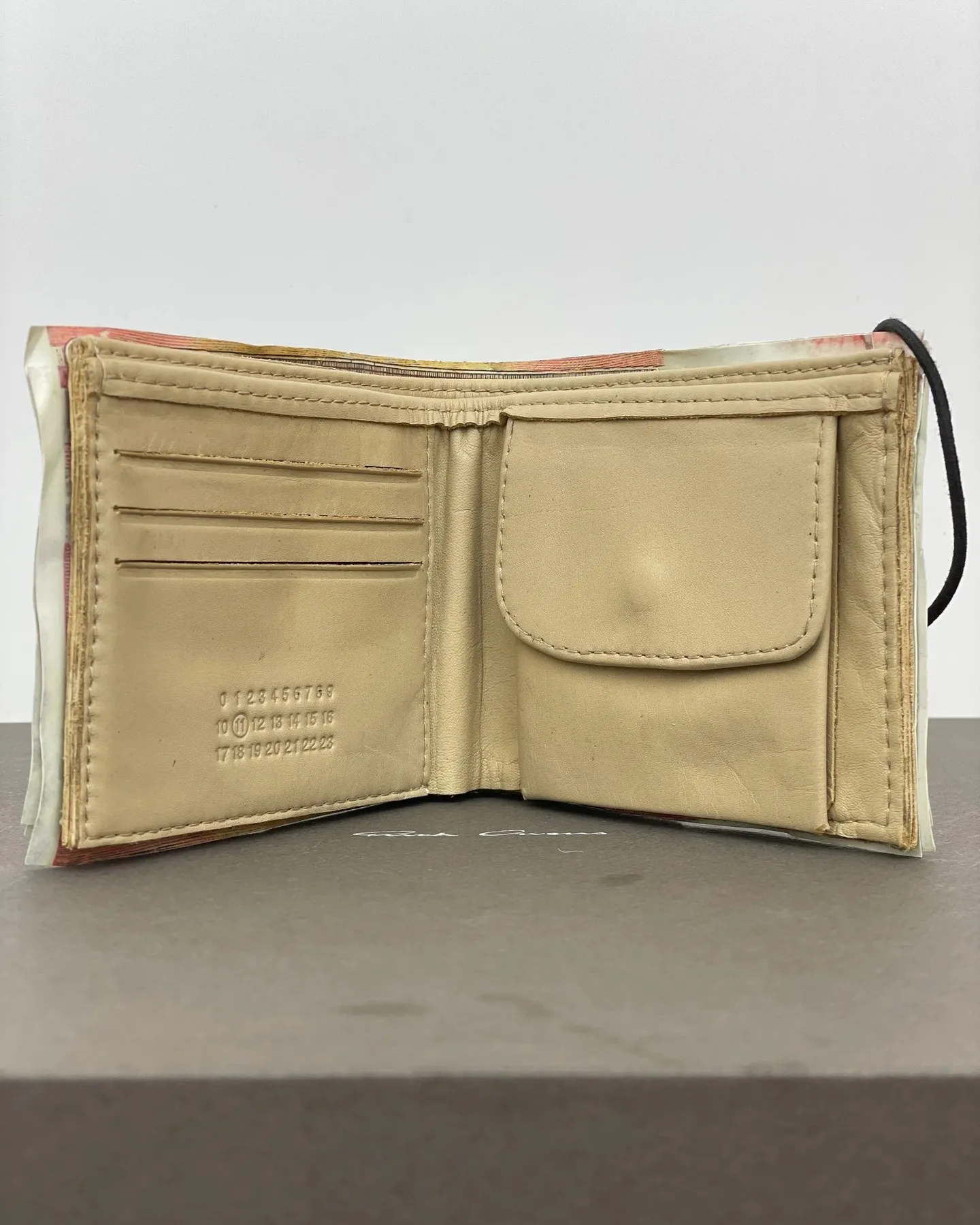 An open leather wallet with cream interior, showing three card slots stamped with the numbers 0 through 23 and 11 circled, and a snap-close coin pocket, wrapped in fake euro notes.