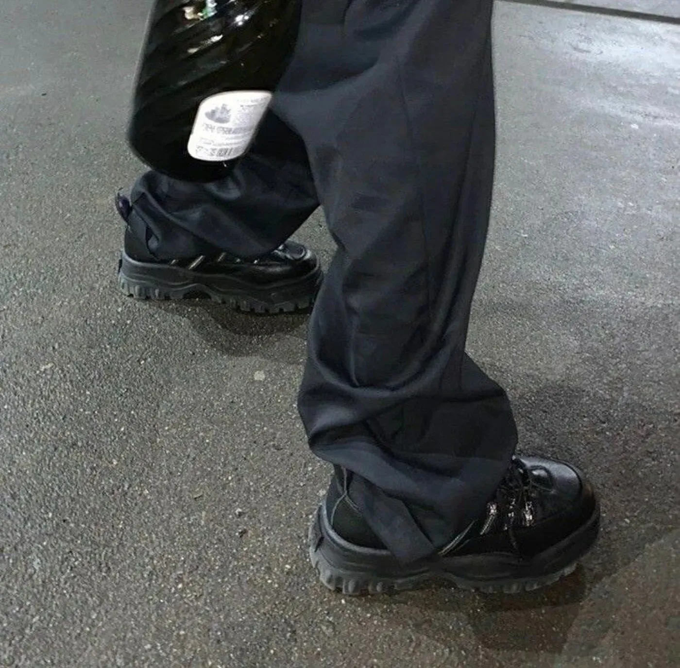Close-up of Ecco2k's feet, wearing black patent leather Eytys Angel Stash sneakers, walking on a wet street.