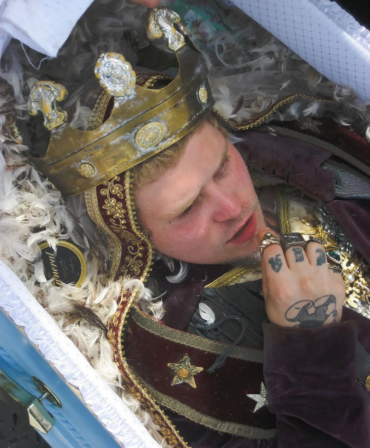 Yung Lean inside the coffin from the 'Forever Yung' video, wearing the gold crown, feathers and fabric around him.
