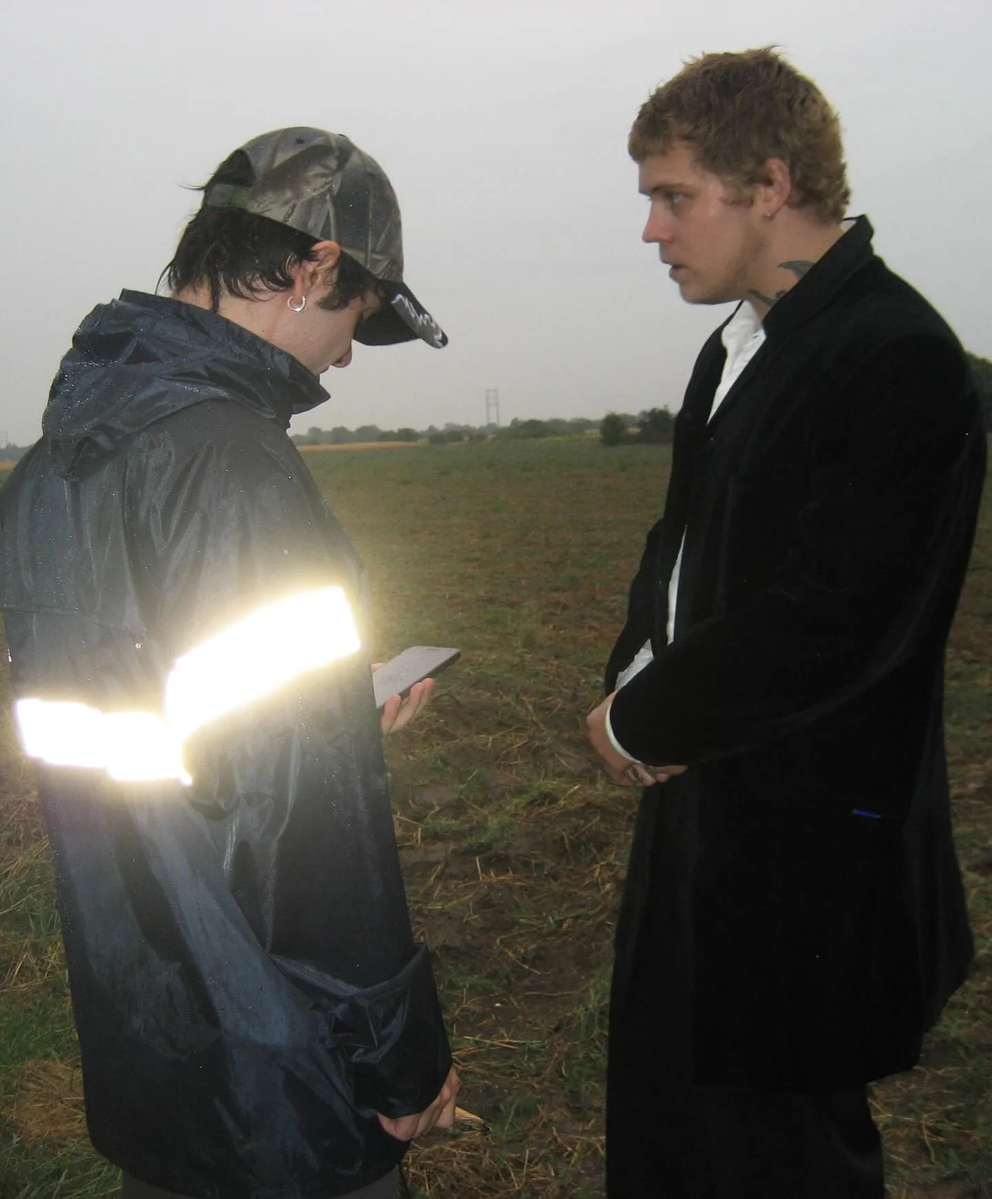 Yung Lean in a long black coat, in conversation with a crew member in a muddy field near the 'Forever Yung' shoot.