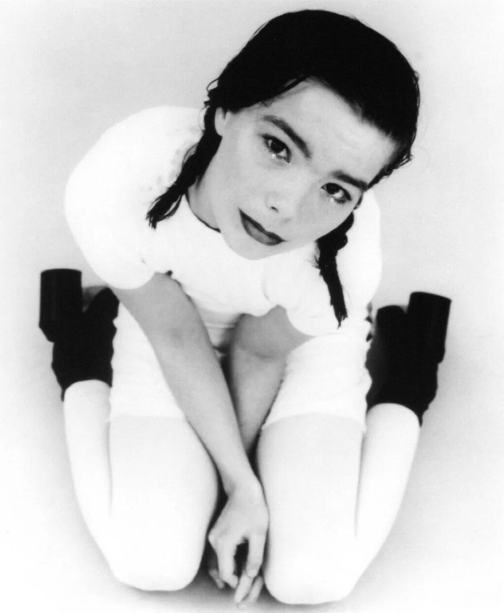 Black and white photograph of Björk seated cross-legged in a white dress, presented as a torn paper cutout collage.