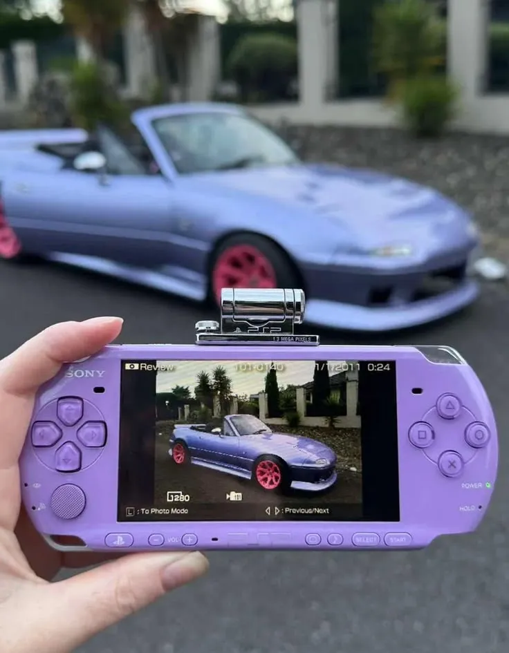 A hand holding a purple PSP with a camera accessory mounted on top, in Review mode, showing a photo of a matching purple roadster with pink wheels parked behind it.