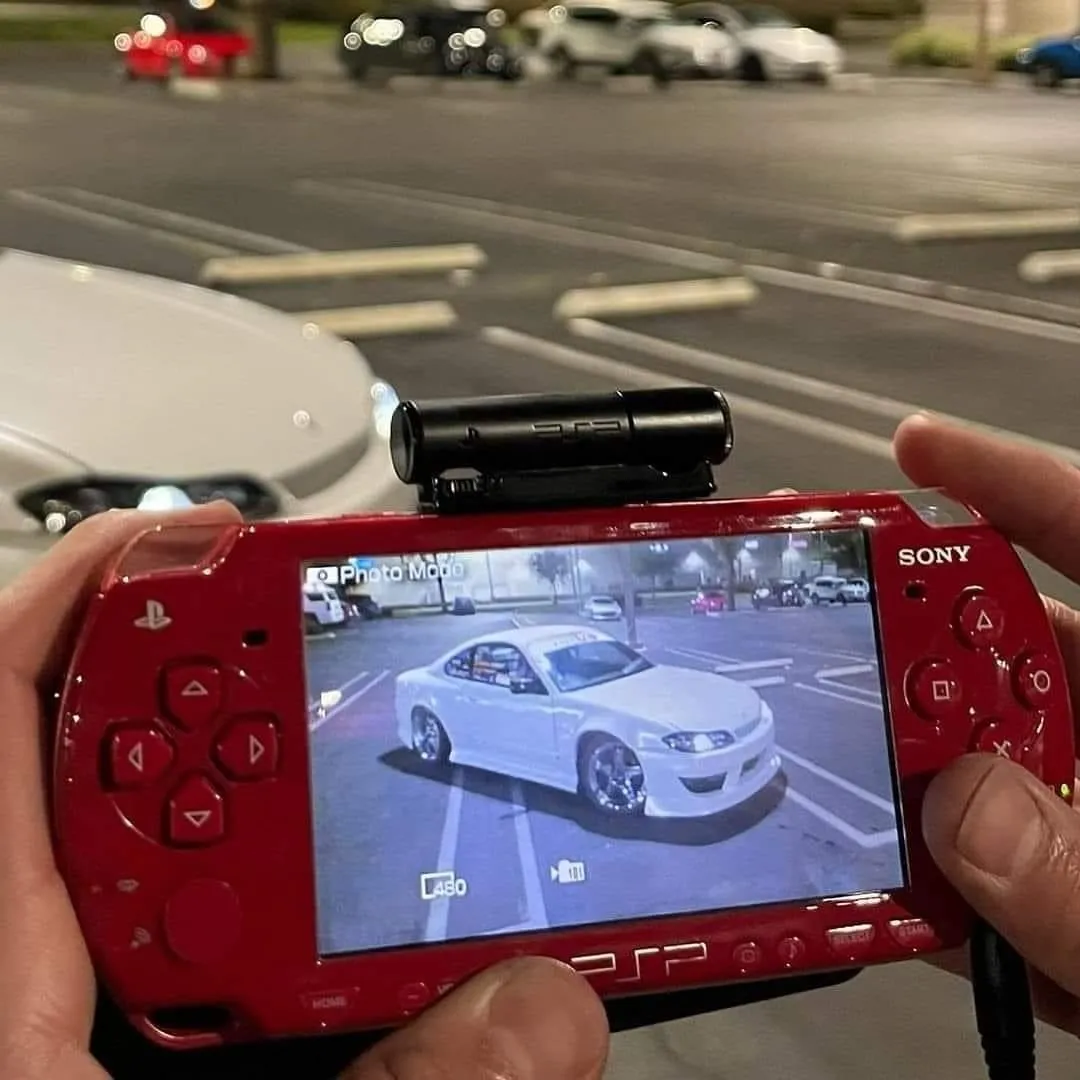 A hand holding a red PSP-1000 with a camera accessory mounted on top, in Photo Mode, aimed at a white modified Nissan Silvia S15 in a parking lot at night.