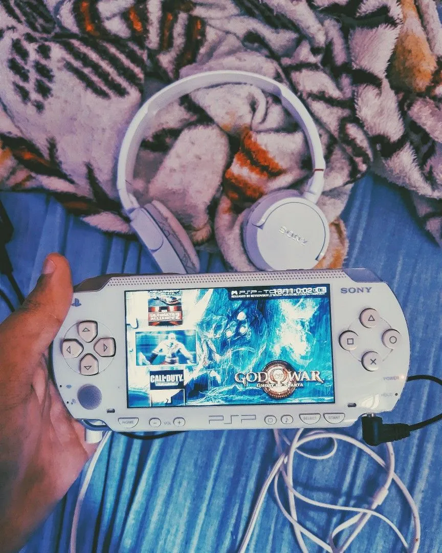 A hand holding a white PSP running God of War Ghost of Sparta, next to a pair of white Sony headphones on a tiger print blanket.