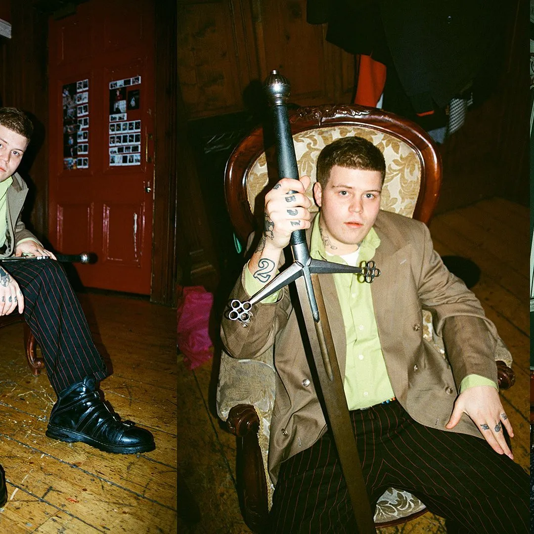 Close-up of Yung Lean's tattooed hand gripping an ornamental silver mace, seated in an antique chair.